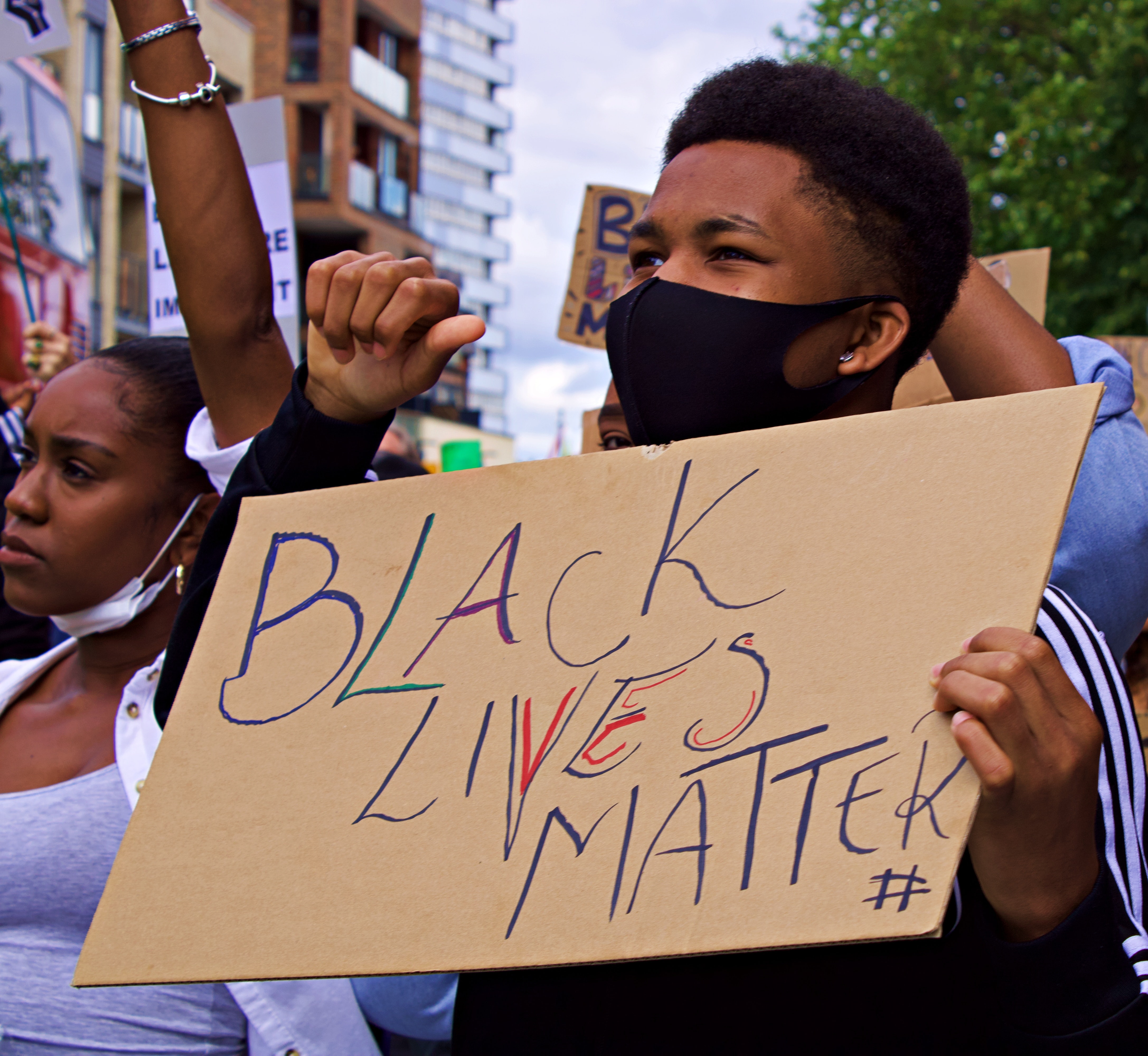 Young Black man wearing a face mask and holding a cardboard sign that says "Black Lives Matter" at an outdoor public demonstration